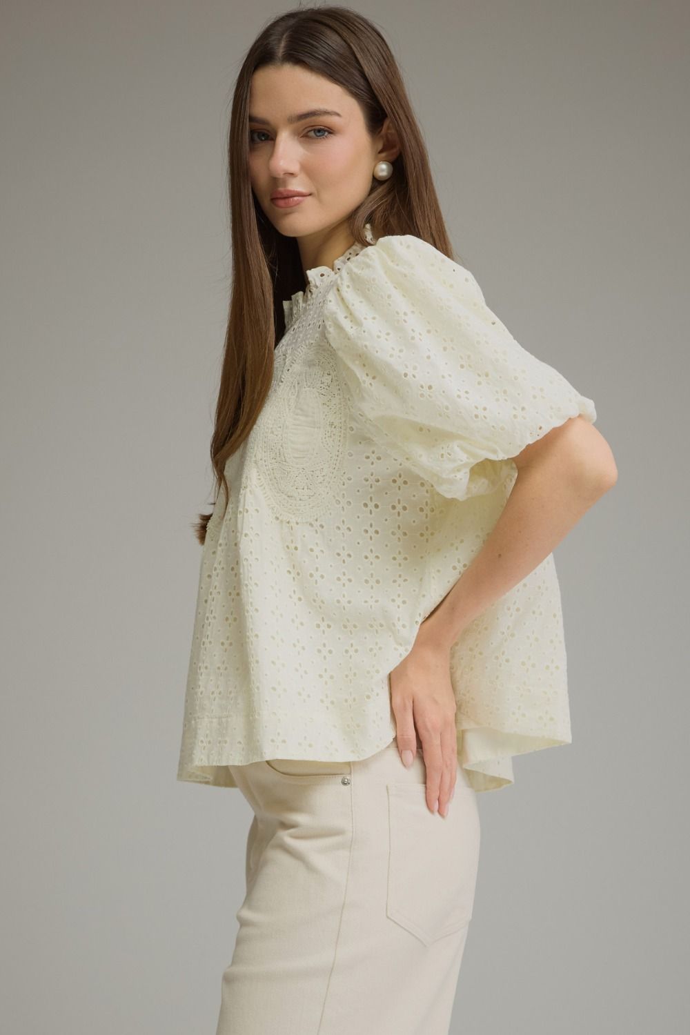 A woman modeling a cream-colored eyelet blouse with voluminous puff sleeves and a high ruffled collar, paired with cream trousers, against a solid gray studio background.