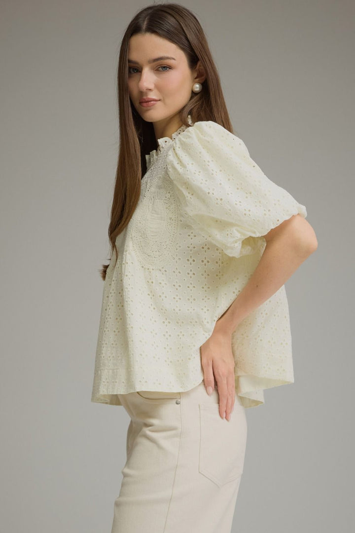 A woman modeling a cream-colored eyelet blouse with voluminous puff sleeves and a high ruffled collar, paired with cream trousers, against a solid gray studio background.