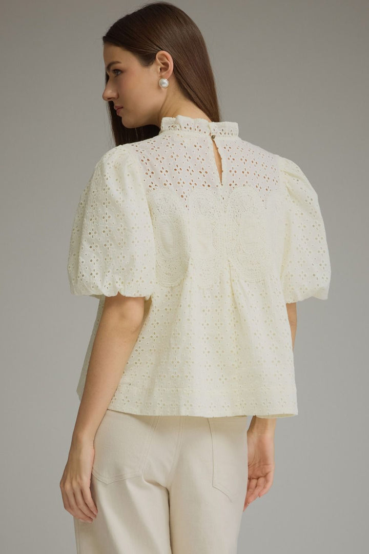 A woman shown from the back wearing a cream-colored eyelet blouse with puff sleeves and a high ruffled collar, paired with matching cream trousers, against a solid gray studio background.