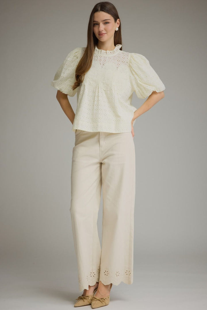 A woman modeling a cream-colored two-piece outfit consisting of a short puff-sleeve eyelet blouse and matching wide-leg trousers, paired with beige pointed-toe flats, against a solid gray studio background.