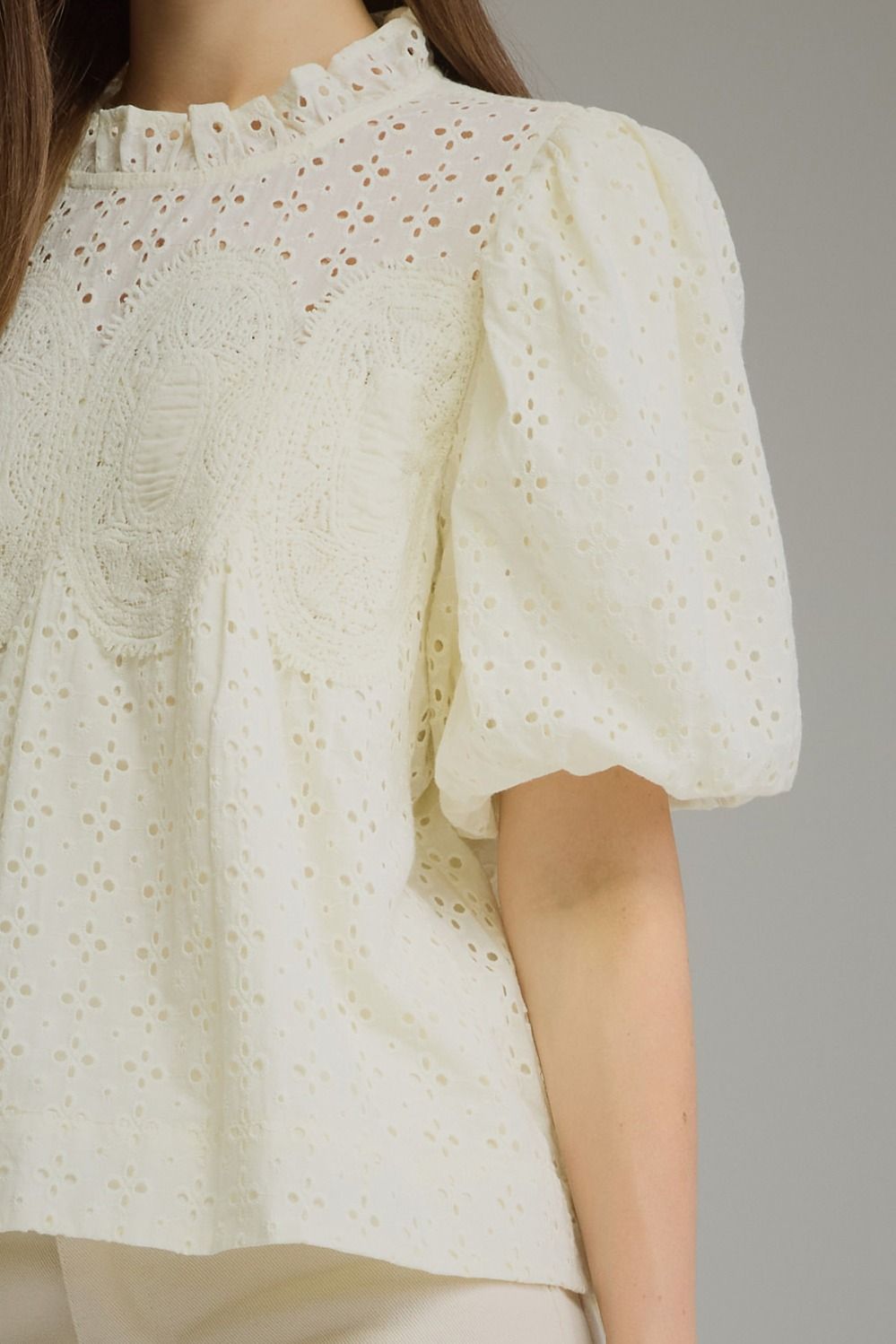 A close-up of a woman wearing a cream-colored eyelet blouse with puff sleeves, a ruffled high neck, and embroidered detailing on the chest.