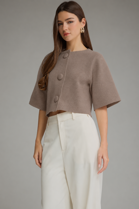 A woman modeling a cropped taupe-brown short-sleeve top with large decorative buttons down the front, paired with high-waisted off-white trousers, against a solid gray studio background.