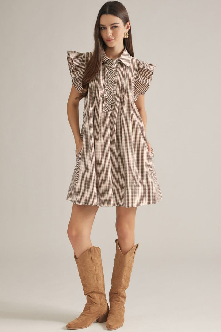 A woman modeling a brown and beige gingham mini dress with ruffled cap sleeves and a smocked bodice, paired with tan knee-high boots, against a neutral studio background.