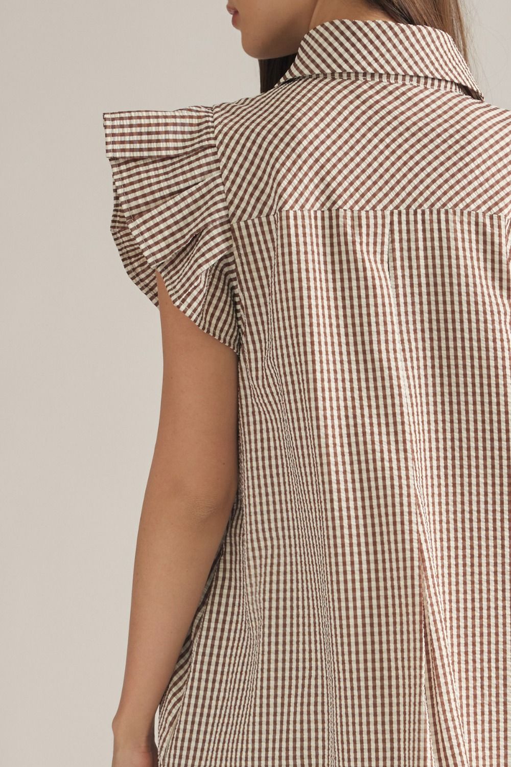 Close-up back view of a model wearing a brown and beige gingham shirt dress with ruffled cap sleeves and a collar, against a light beige background.