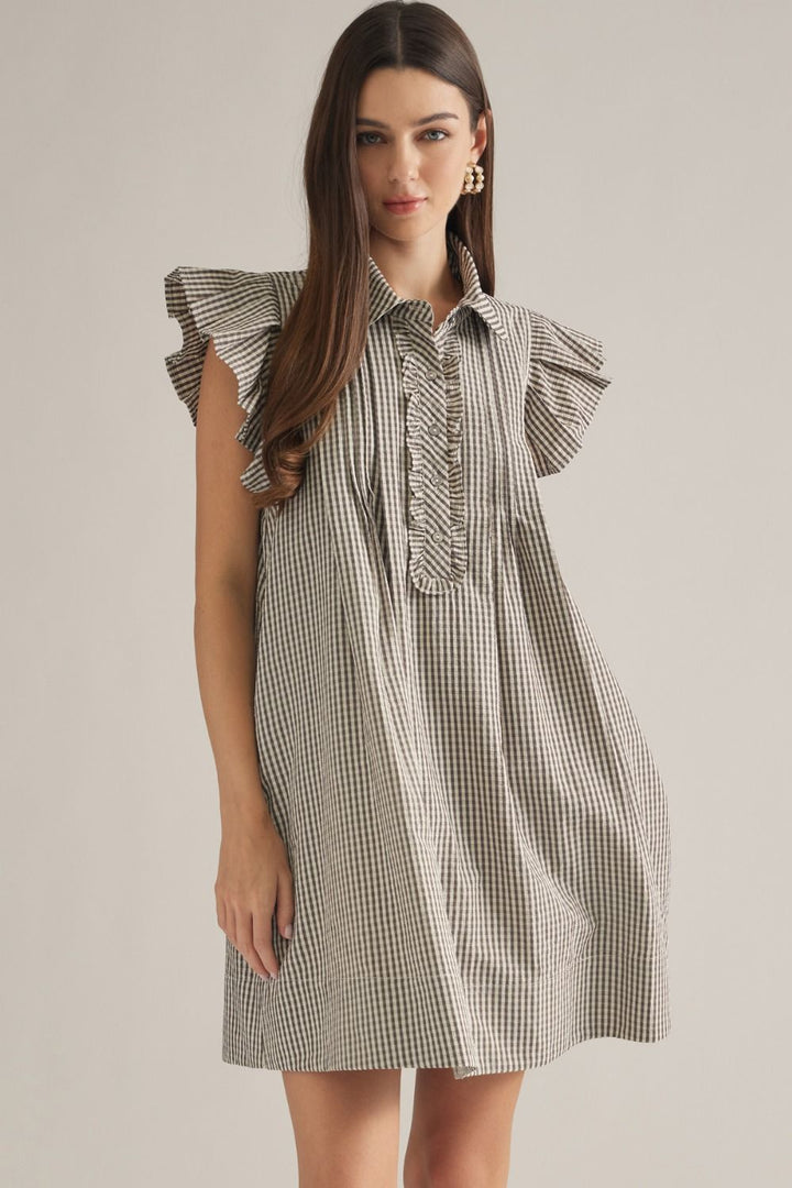 A woman modeling a short gingham dress with ruffled cap sleeves and a buttoned collar, photographed against a plain light beige studio background.