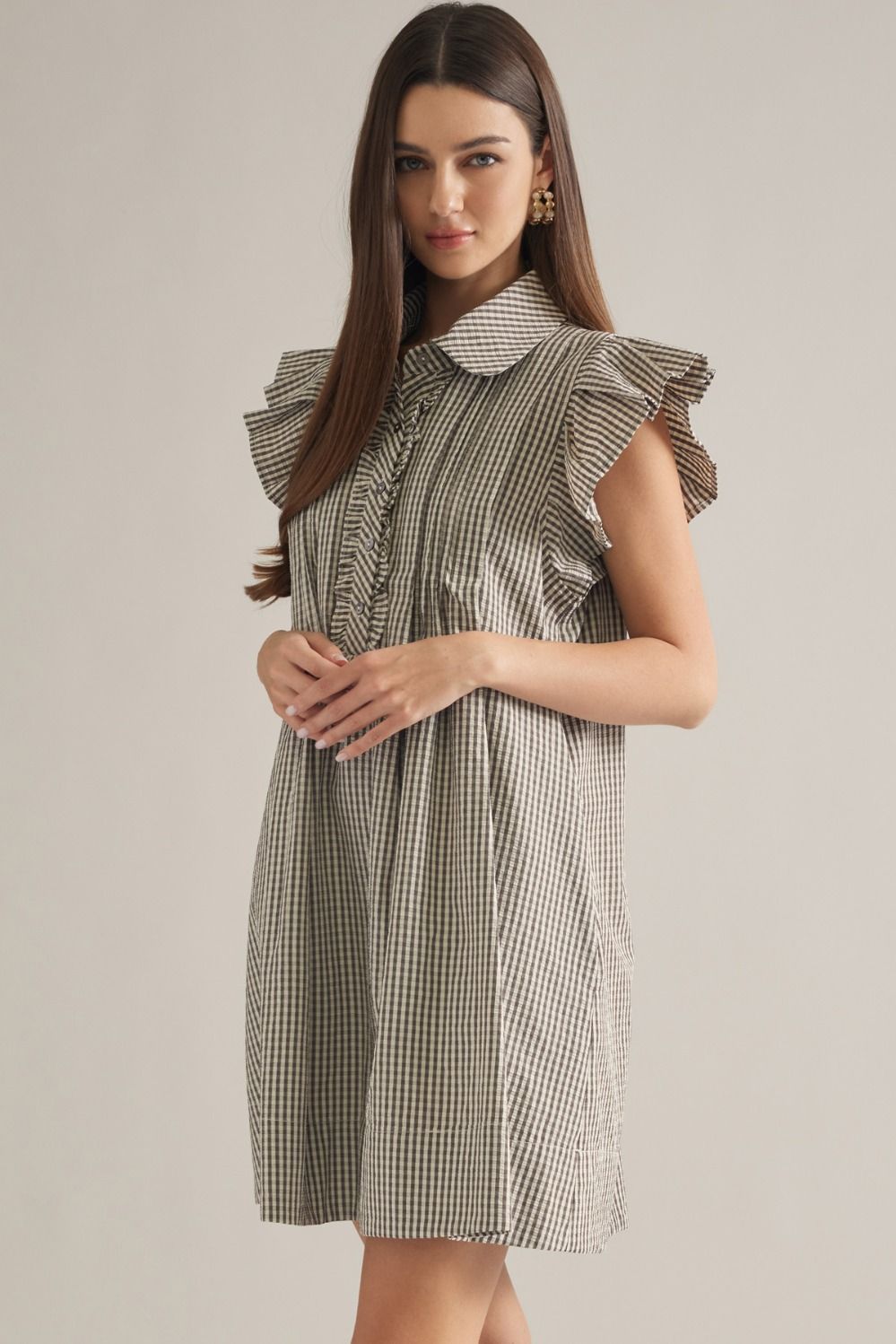 A woman modeling a sleeveless, high-neck dress with a brown and white gingham pattern and ruffled details on the shoulders and collar, set against a plain light gray studio background.