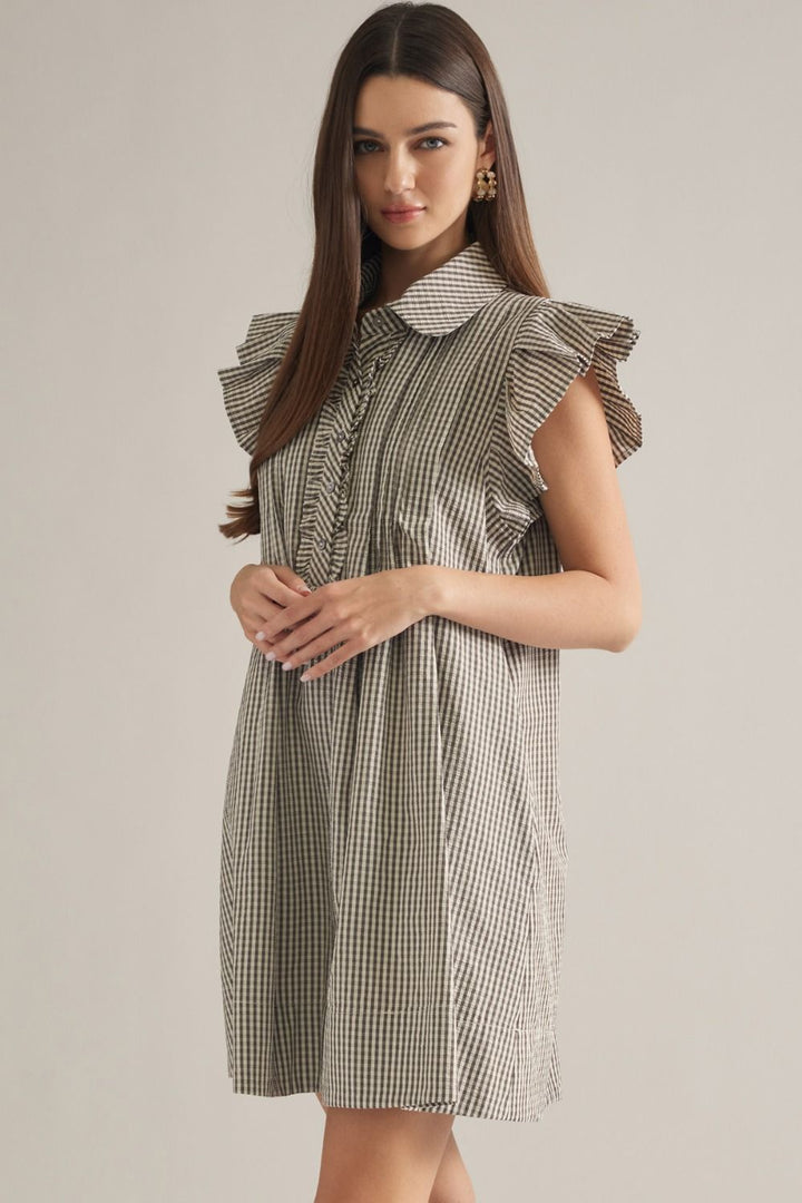 A woman modeling a sleeveless, high-neck dress with a brown and white gingham pattern and ruffled details on the shoulders and collar, set against a plain light gray studio background.