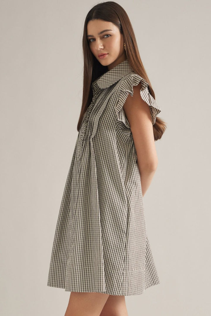 A woman modeling a sleeveless mini dress with a small-scale black and white gingham pattern, ruffled cap sleeves, and a high collar against a plain light beige studio background.