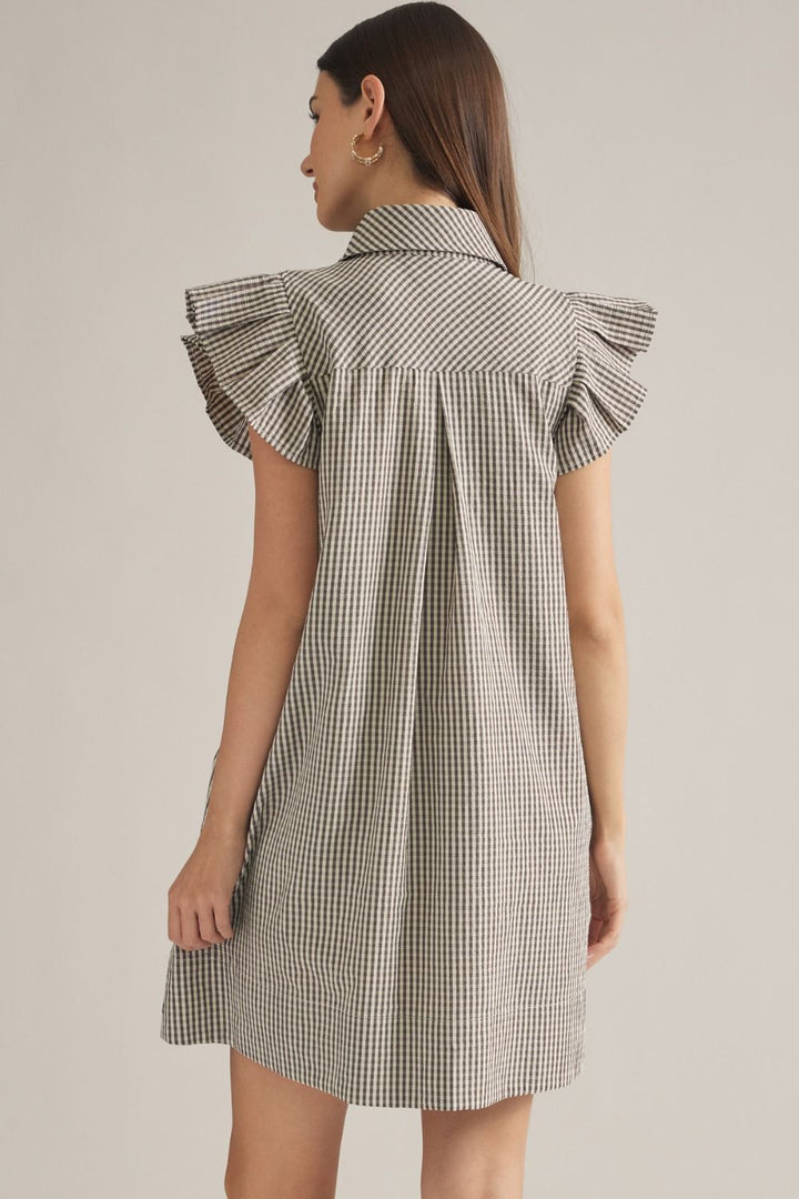 A woman shown from the back wearing a short-sleeve gingham dress with ruffled shoulders, photographed against a plain light beige studio background.