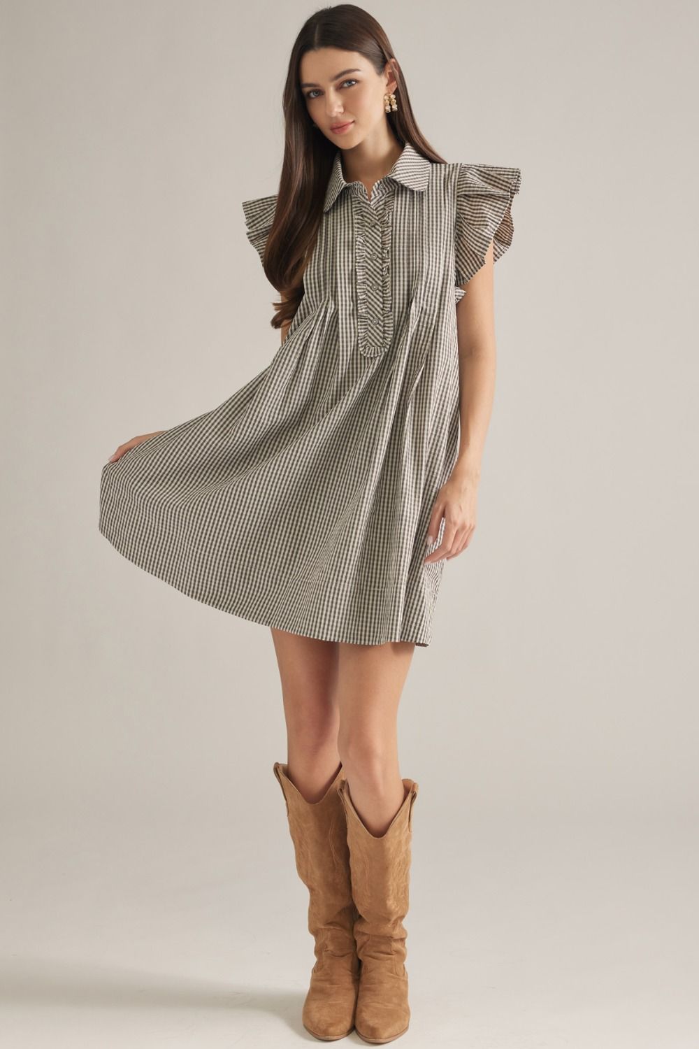 A woman modeling a short-sleeve collared dress with a fine black and white gingham pattern, ruffled shoulders, and a gathered front, paired with tan cowboy boots against a light gray studio background.