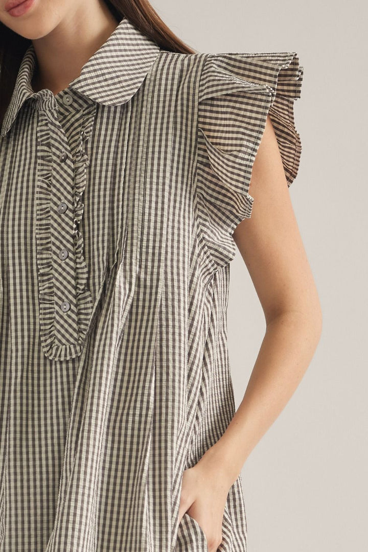 Close-up of a woman wearing a sleeveless gingham dress with ruffled cap collar, ruffled shoulder details, and a button placket, against a plain light beige background.