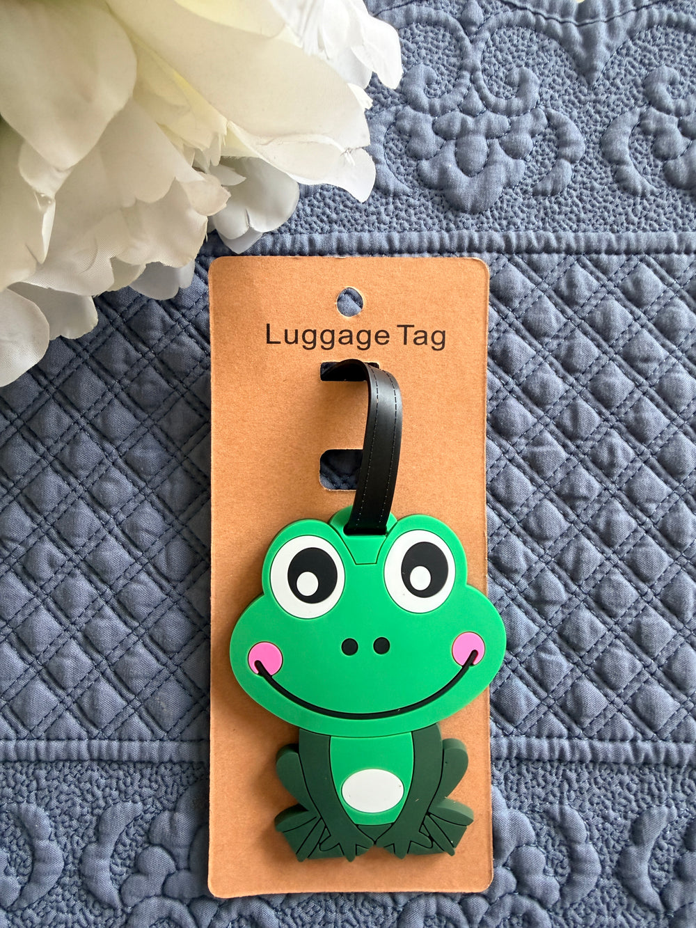Green frog-shaped luggage tag on a brown card with 'Luggage Tag' text, against a textured gray background.