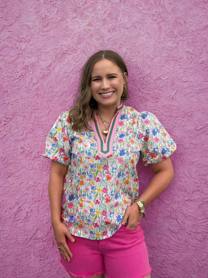 Woman wearing a colorful floral blouse and pink shorts against a pink textured wall.