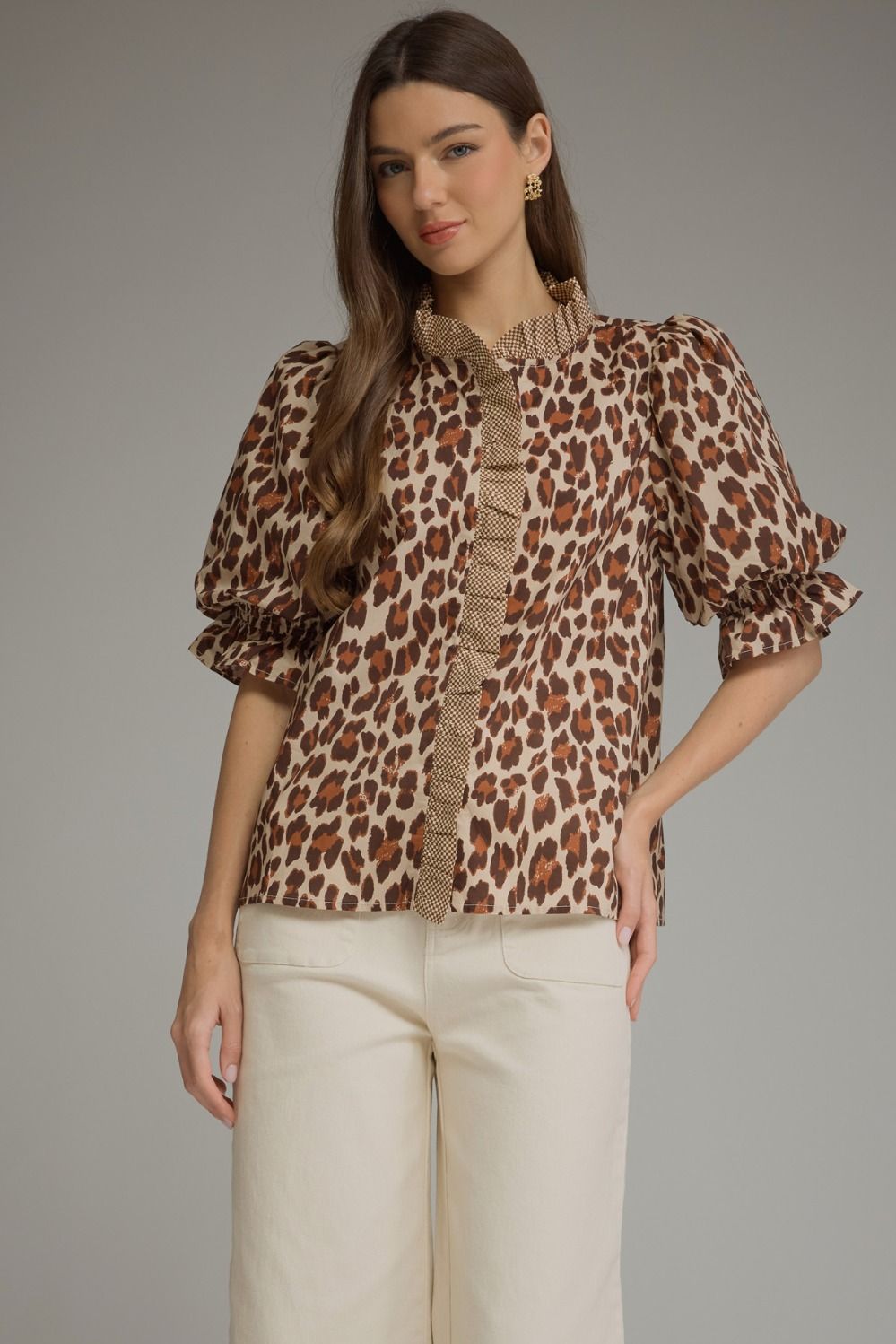 A woman modeling a leopard-print blouse with a textured ruffle collar and puff sleeves, paired with cream-colored pants, against a solid gray studio background.