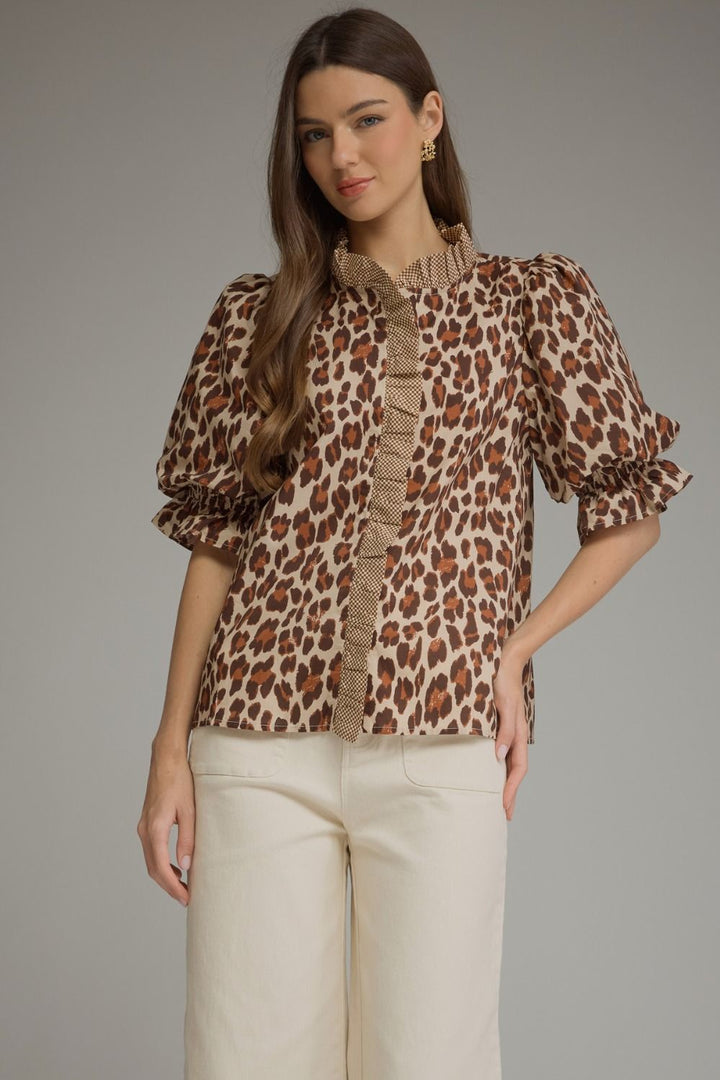 A woman modeling a leopard-print blouse with a textured ruffle collar and puff sleeves, paired with cream-colored pants, against a solid gray studio background.