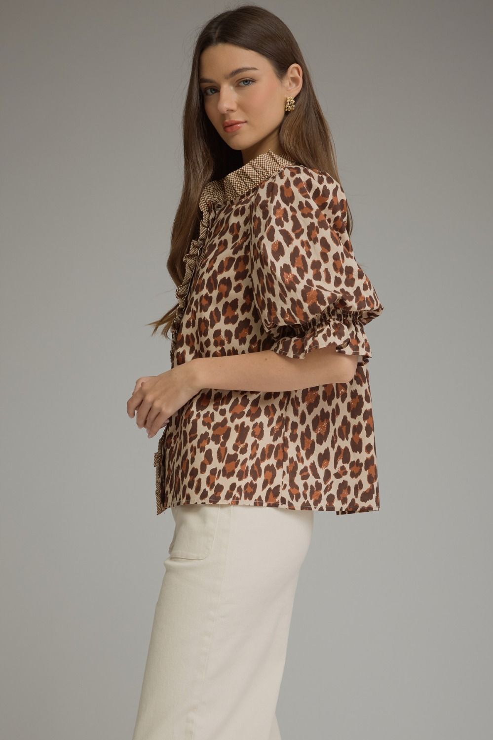 A woman modeling a leopard-print blouse with ruffled details on the collar and sleeves, paired with white trousers, against a solid gray studio background.