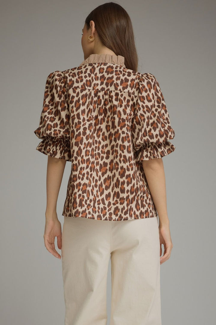 A woman shown from the back wearing a leopard-print blouse with ruffled short sleeves and a pleated high neckline, paired with light beige pants, against a plain gray studio background.