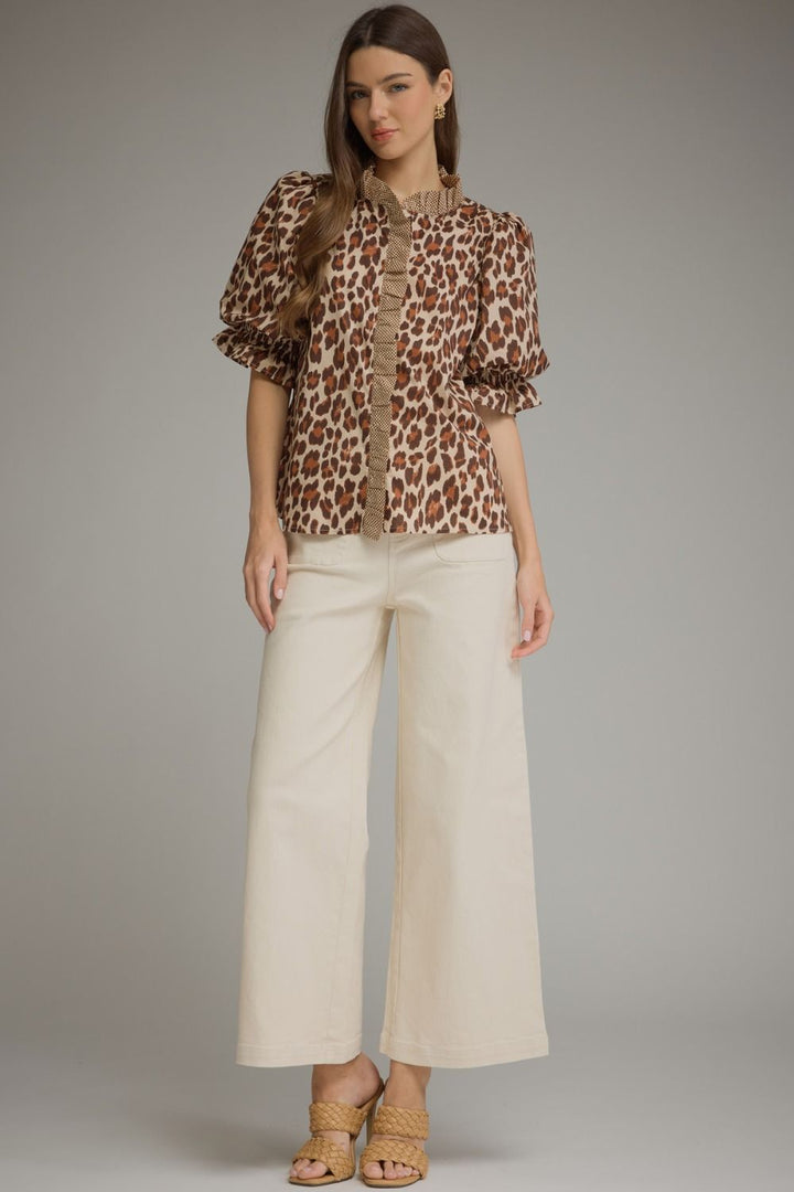 A woman modeling a brown and beige leopard-print short-sleeve blouse with ruffled trim, paired with cream wide-leg cropped pants and tan strappy heels, against a neutral gray studio background.