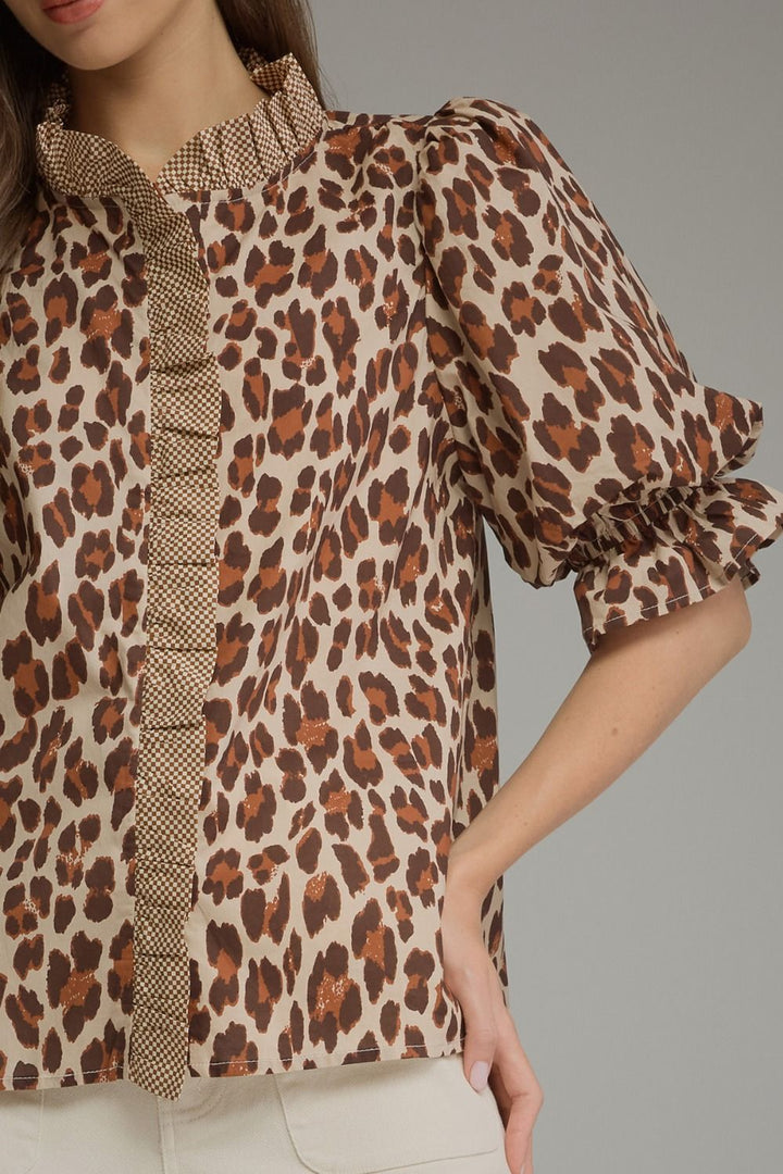 A close-up of a woman wearing a leopard print blouse with ruffle trim on the collar, front placket, and sleeve cuffs, paired with light beige pants against a neutral gray background.