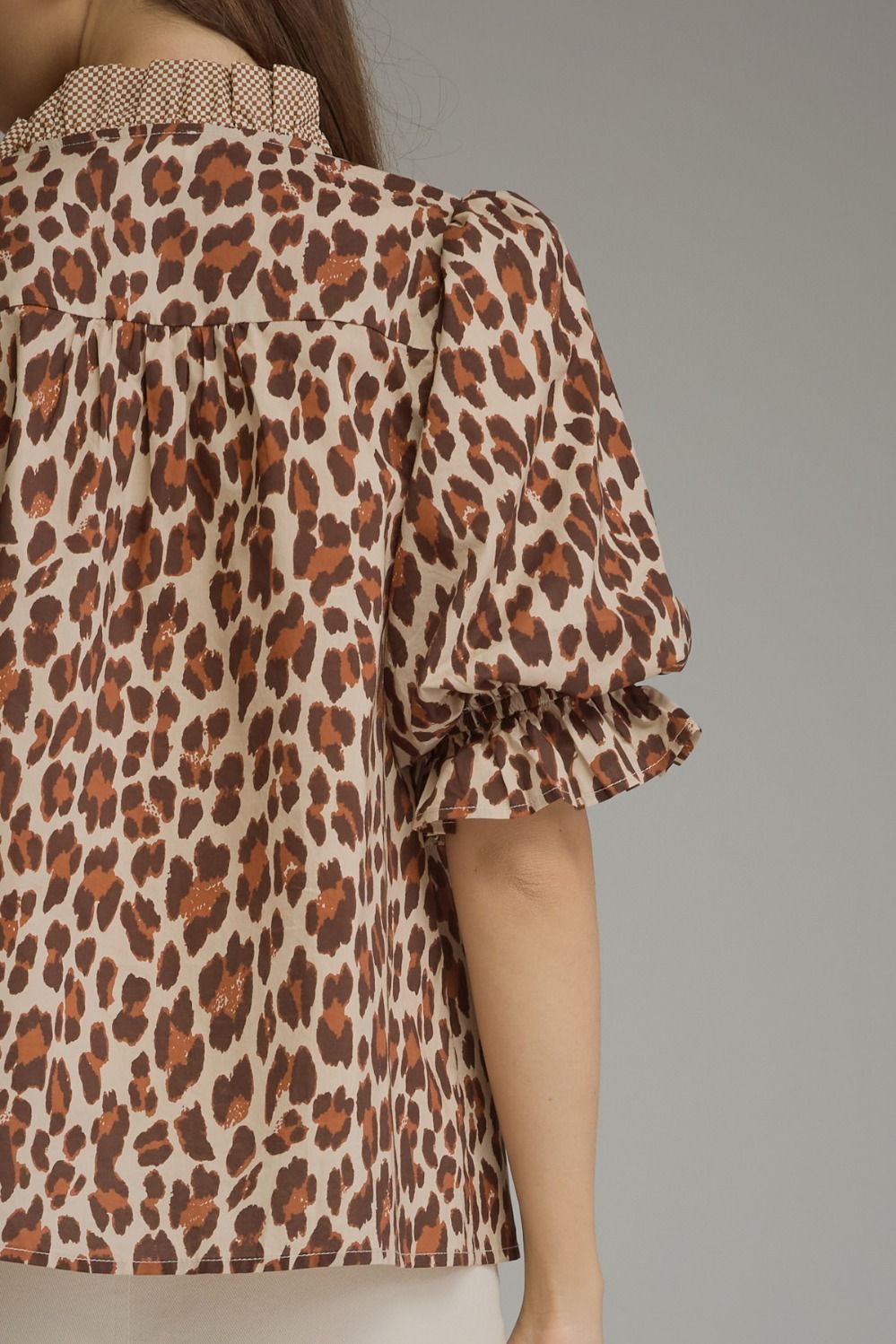A close-up of a model wearing a leopard print blouse with puff sleeves and a ruffled collar, shown from the back against a solid gray background.
