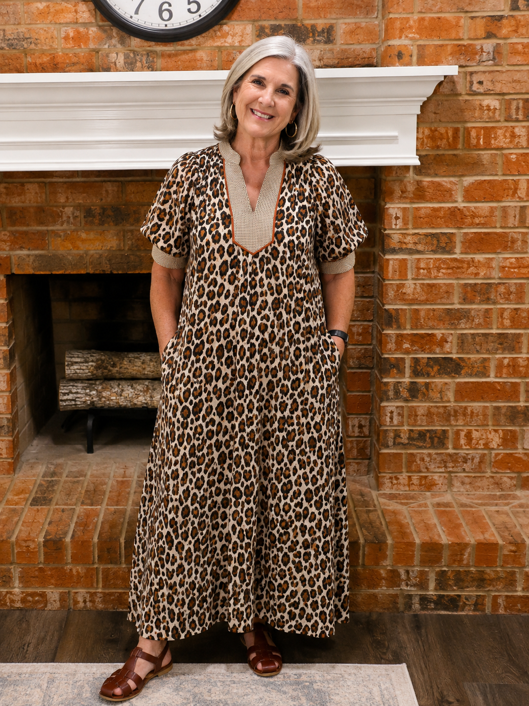 A woman with short gray hair wearing a brown and black leopard print maxi dress with beige trim, standing in front of a brick fireplace.