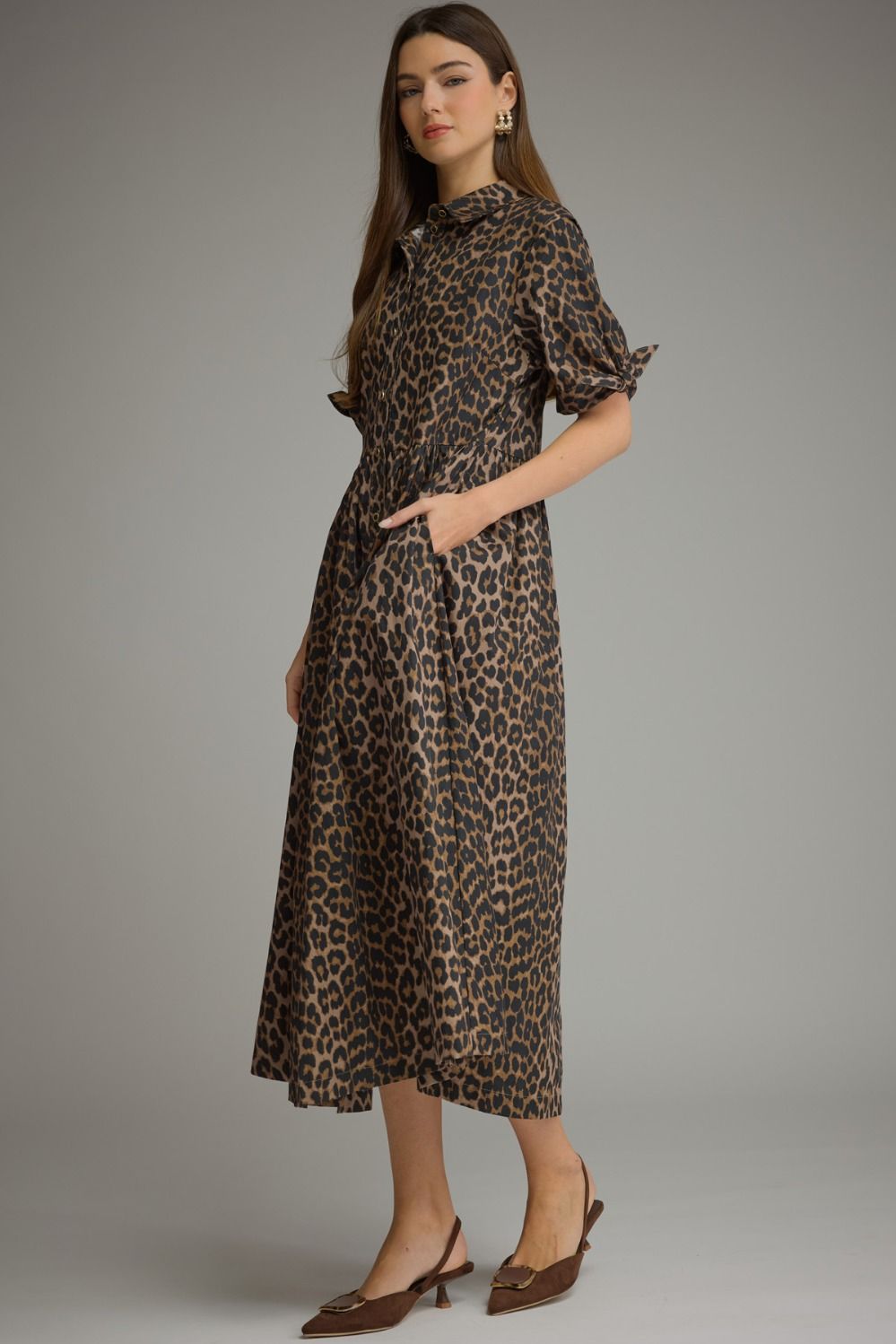 A woman modeling a leopard-print midi dress with puff sleeves and side pockets, paired with brown pointed-toe heels, against a solid gray studio background.