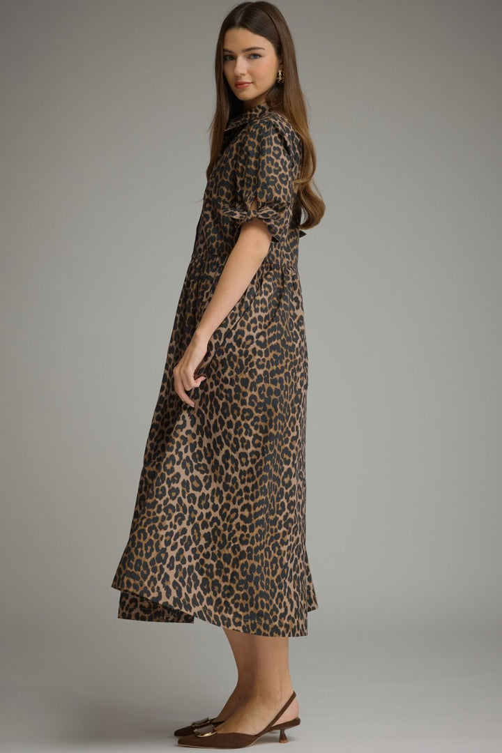 A woman modeling a leopard-print midi dress with short puff sleeves, a high collar, and a flared hem, photographed against a plain gray studio background.