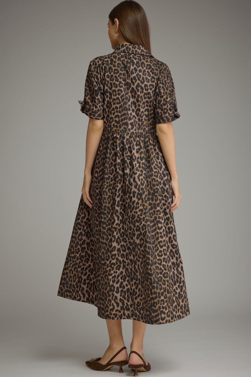 A woman viewed from the back wearing a leopard-print midi dress with short ruffled sleeves and brown heeled sandals, photographed against a plain gray studio background.