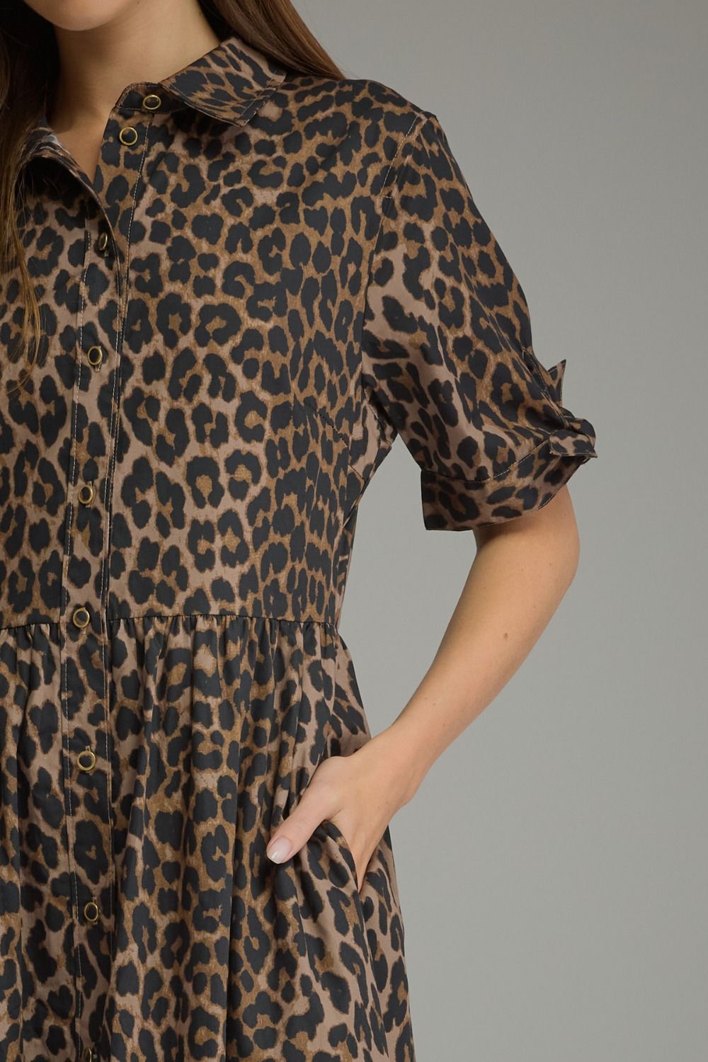 A close-up of a woman wearing a leopard print shirt dress with short puff sleeves, a button-down front, and side pockets, set against a solid gray background.