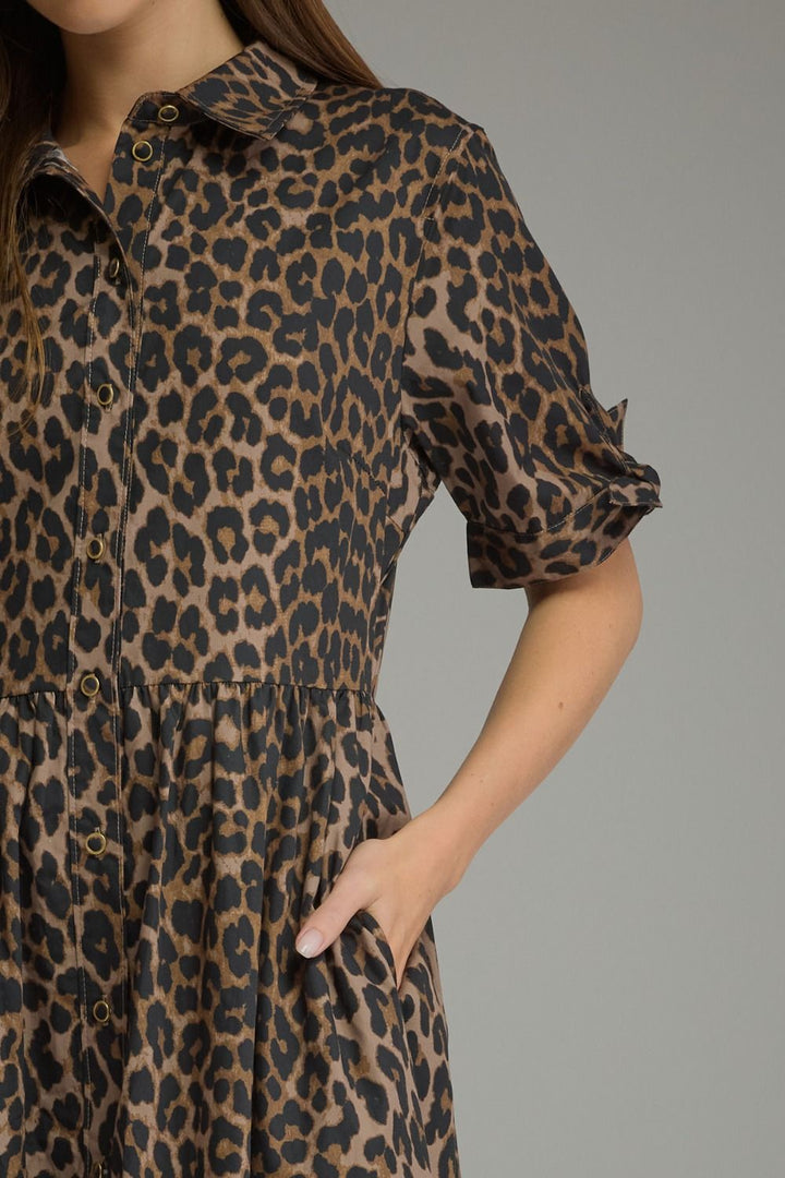 A close-up of a woman wearing a leopard print shirt dress with short puff sleeves, a button-down front, and side pockets, set against a solid gray background.