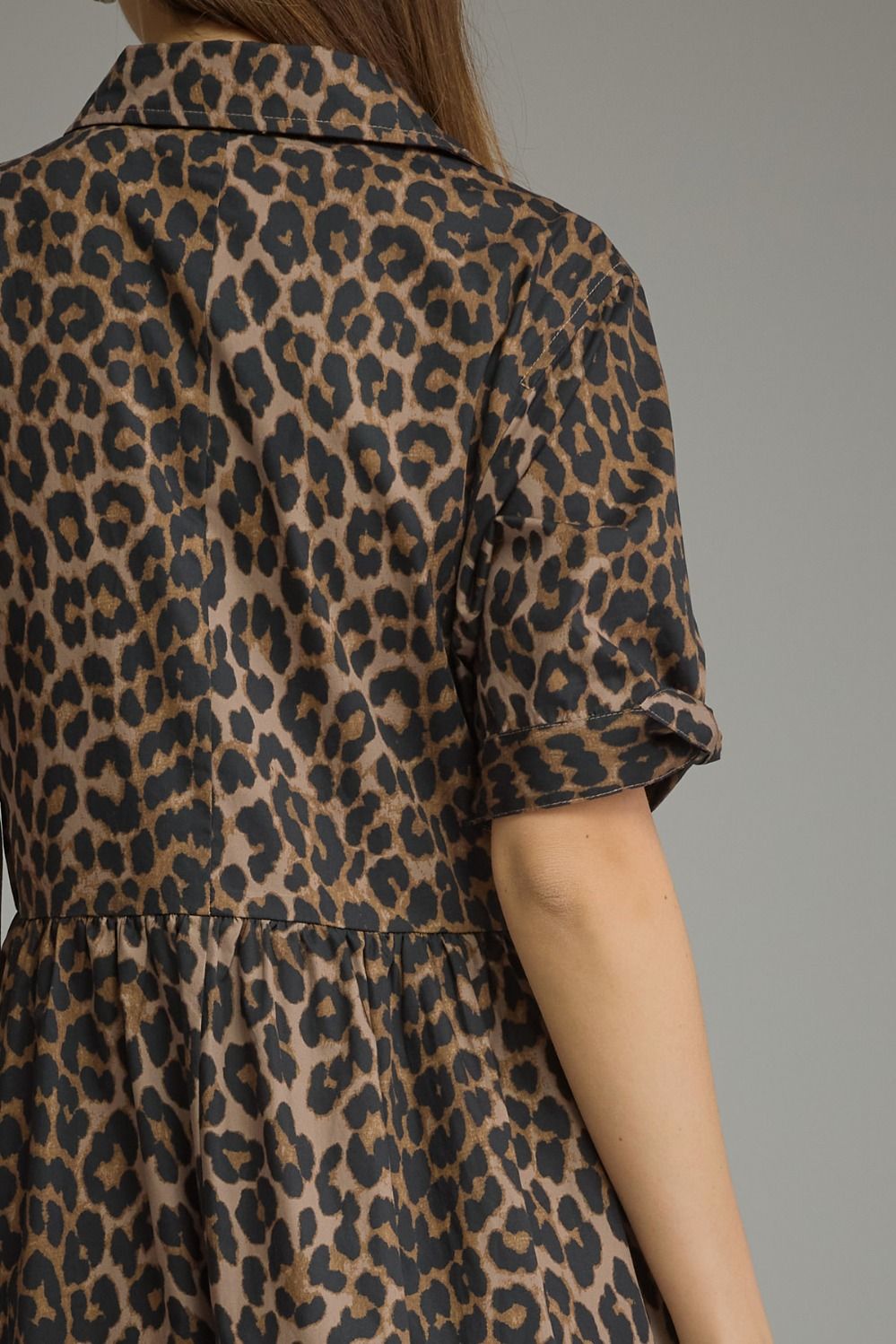 Close-up back view of a person wearing a leopard-print dress with short sleeves and a gathered waist, against a plain gray background.