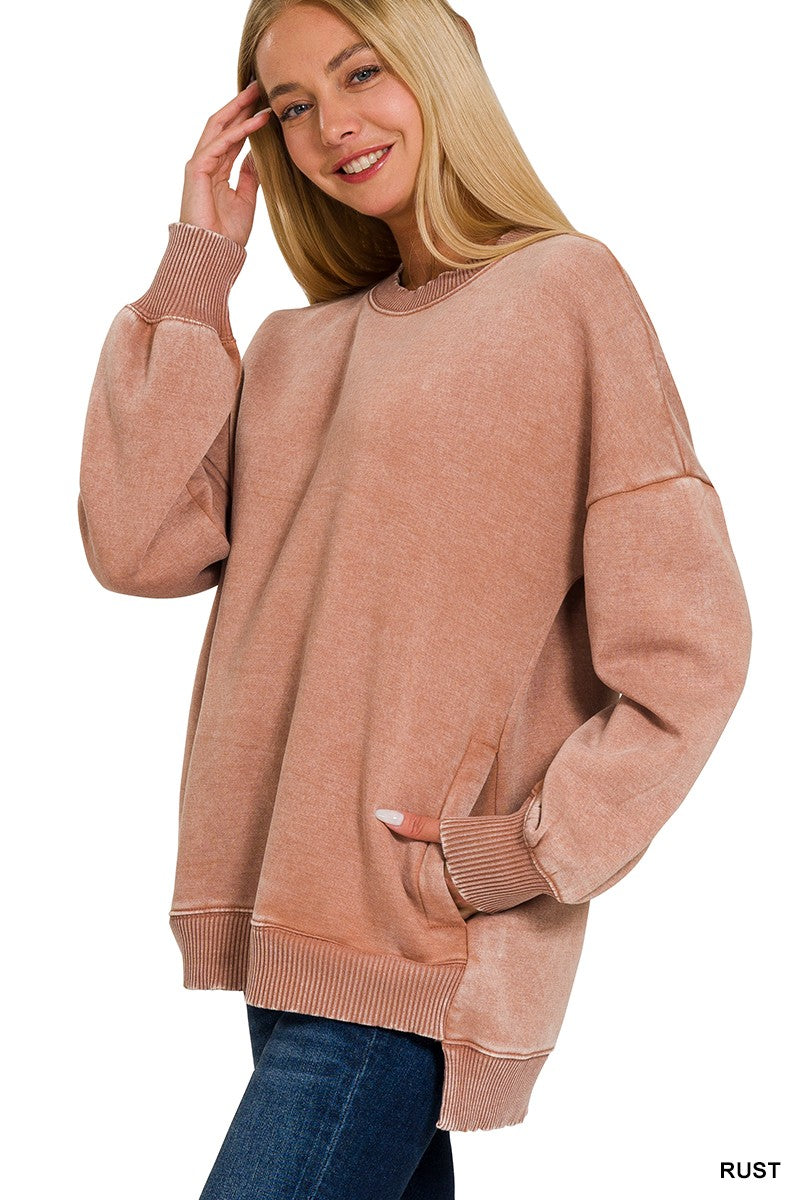 A woman wearing a dusty rose or rust-colored oversized crewneck sweatshirt with long sleeves and ribbed cuffs, hem, and collar, paired with blue jeans against a white background.