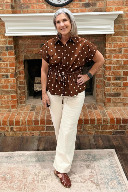 A woman with gray hair stands in front of a brick fireplace wearing a brown short-sleeve button-up shirt with white polka dots and cream-colored pants.