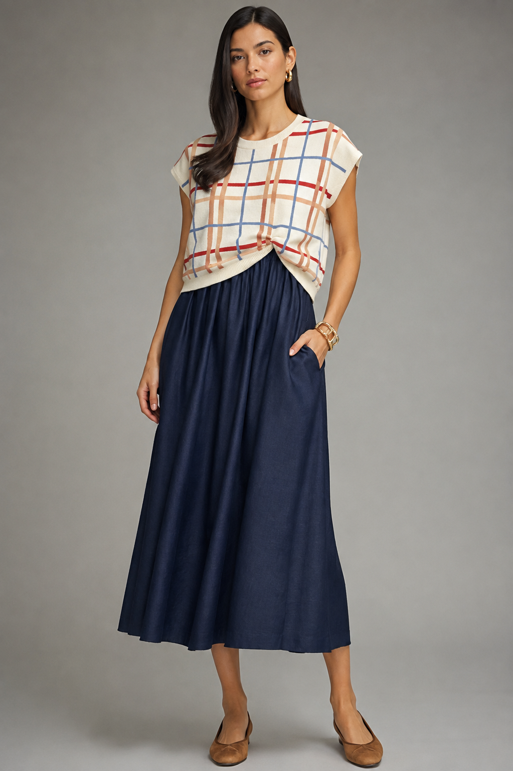 A woman modeling a sleeveless knit top with a cream base and red, blue, and orange plaid pattern, paired with a navy midi skirt and tan flats, against a solid gray studio background.
