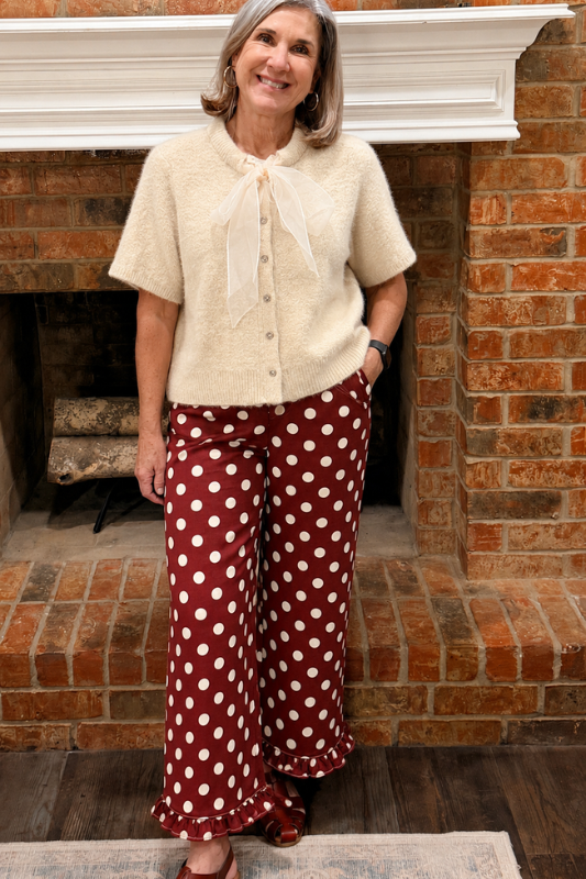 A woman stands in front of a brick fireplace wearing a cream short-sleeve fuzzy button-front top with a sheer bow tie detail and burgundy wide-leg pants with white polka dots.
