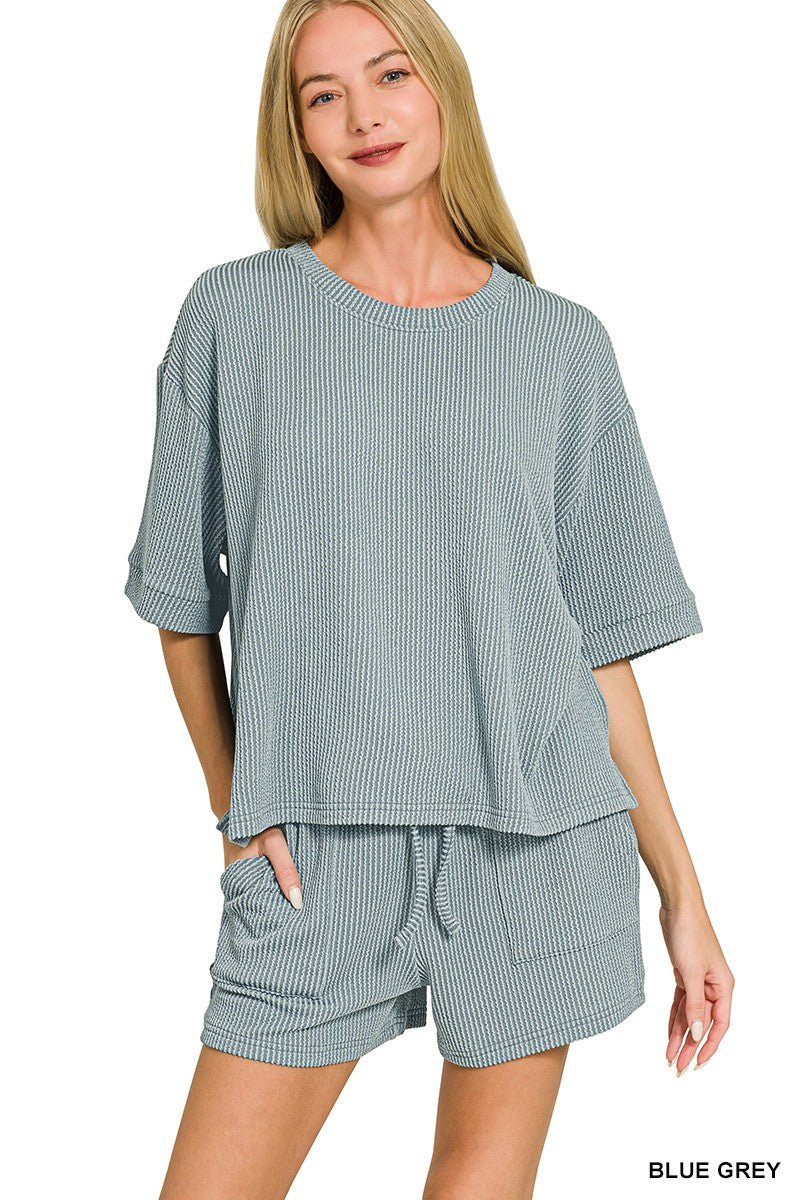 A woman modeling a matching two-piece loungewear set consisting of a short-sleeve top and shorts in a blue-grey color with thin white vertical pinstripes, set against a plain white background.