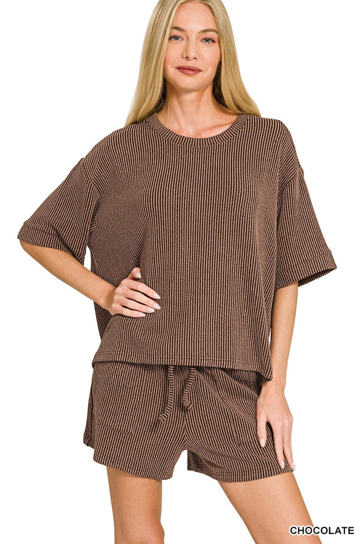 A woman modeling a matching brown ribbed knit lounge set consisting of a short-sleeve top and drawstring shorts, against a plain white background.