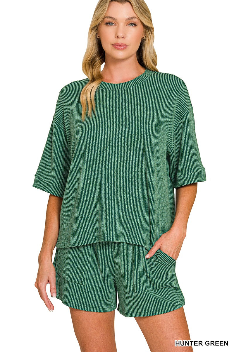 A woman modeling a matching green ribbed knit lounge set consisting of a short-sleeve top and shorts, against a plain white background.