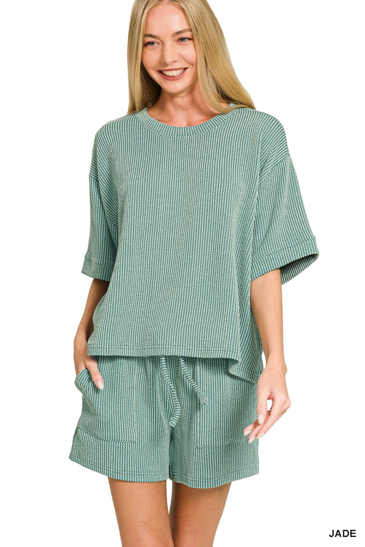 A woman modeling a matching two-piece lounge set in a muted sage green color with a fine vertical stripe pattern. The set includes a short-sleeve top and drawstring shorts, shown against a plain white background.