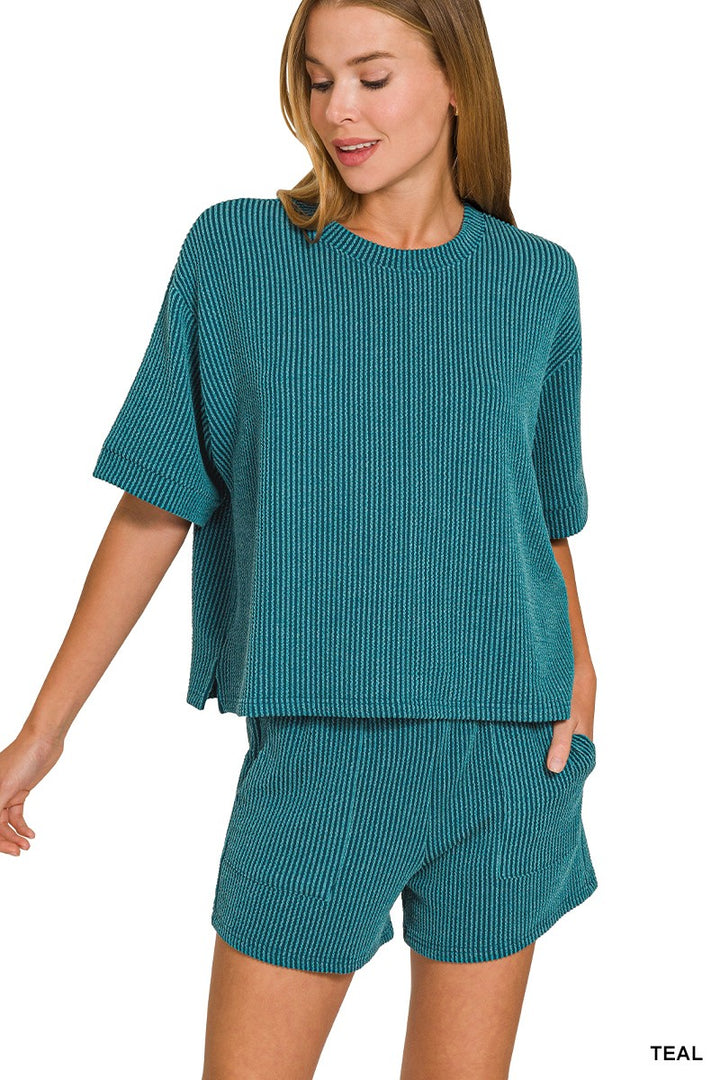 A woman modeling a matching teal ribbed knit lounge set consisting of a short-sleeve top and shorts, shown against a plain white background.