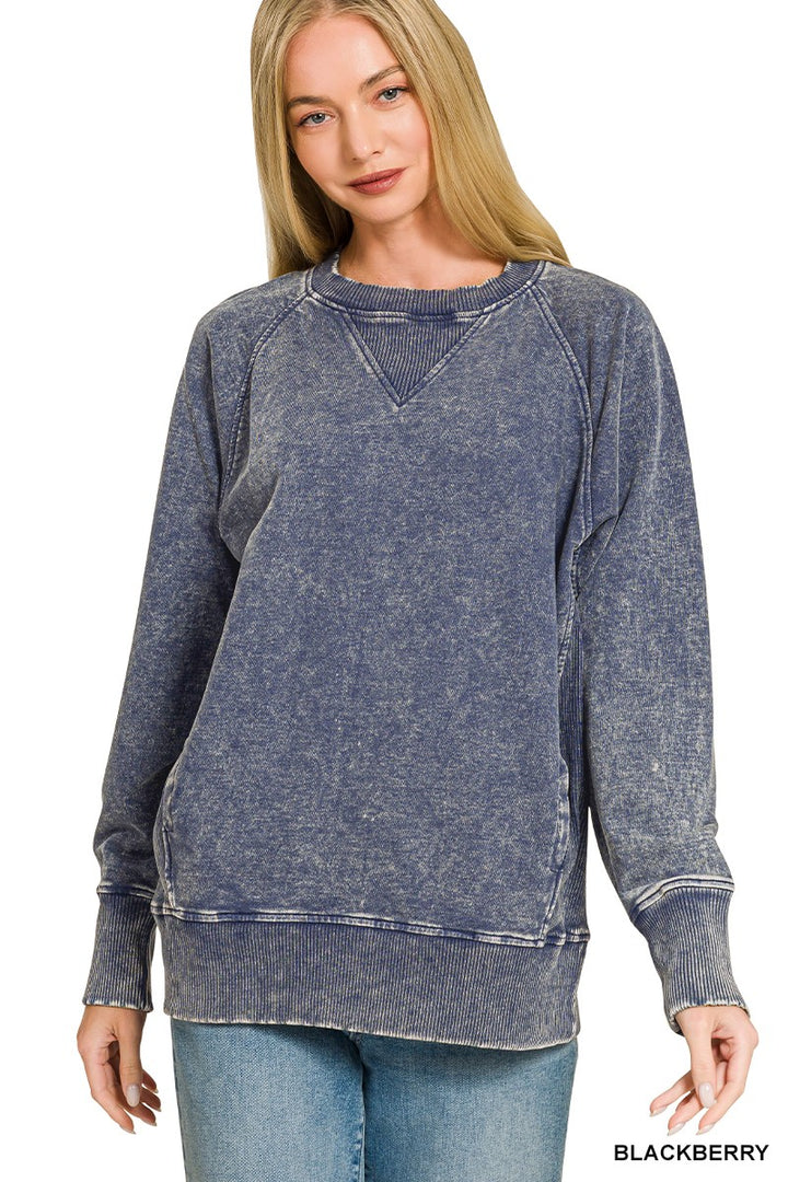 A woman modeling a washed slate blue crewneck sweatshirt with a V-neckline, paired with blue jeans, against a plain white background. The word &