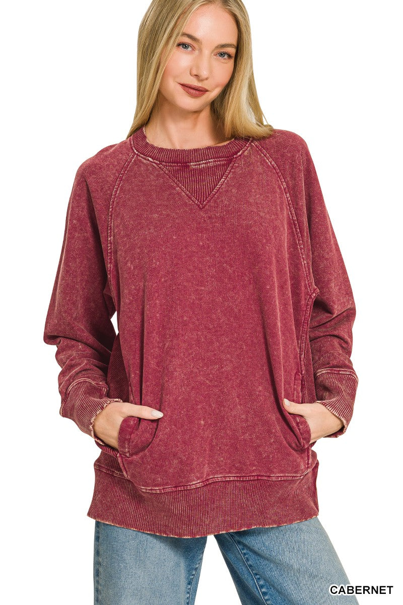 A woman wearing a long-sleeve, oversized crewneck sweatshirt in a washed cabernet red color, paired with blue jeans, against a plain white background.