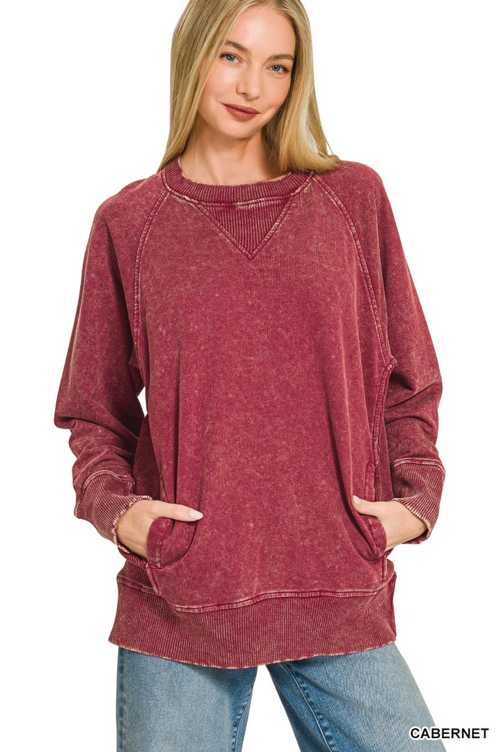 A woman wearing a long-sleeve, oversized crewneck sweatshirt in a washed cabernet red color, paired with blue jeans, against a plain white background.