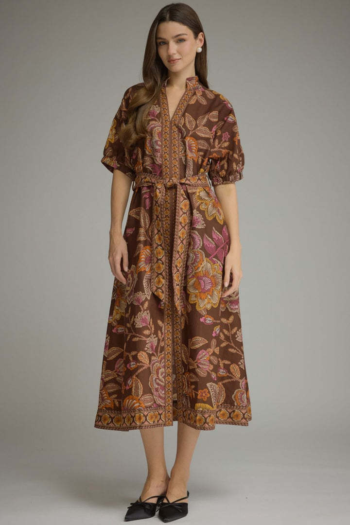 A woman modeling a brown midi-length wrap dress with a colorful floral and paisley print in pink, orange, and yellow, paired with black pointed flats, against a neutral gray studio background.
