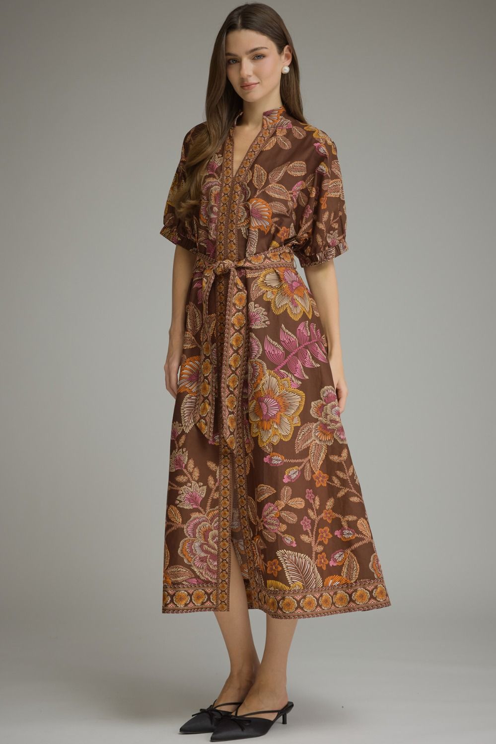 A woman modeling a brown midi wrap dress with a vibrant floral and paisley print in pink, orange, and yellow, paired with black pointed-toe flats, against a solid gray studio background.