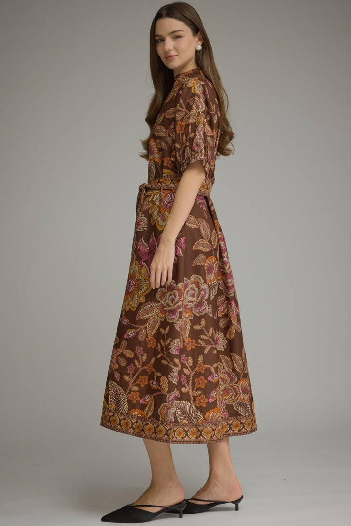 A woman modeling a brown midi dress with a vibrant batik floral pattern in orange, pink, and yellow, paired with black pointed flats, against a solid gray studio background.