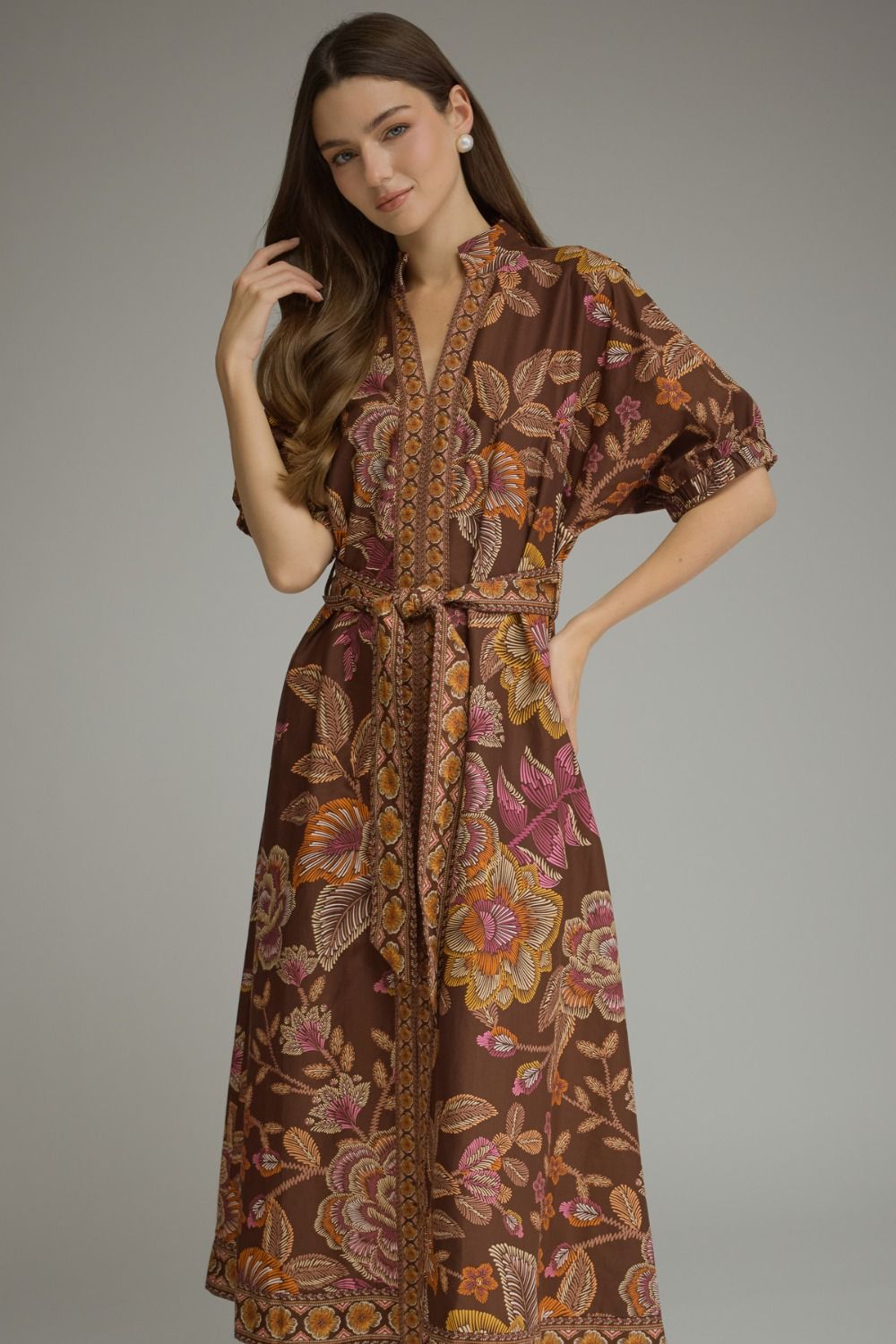 A woman modeling a brown batik maxi dress with a floral pattern in pink, orange, and yellow, featuring a V-neckline, short puff sleeves, and a matching waist tie, set against a plain gray studio background.