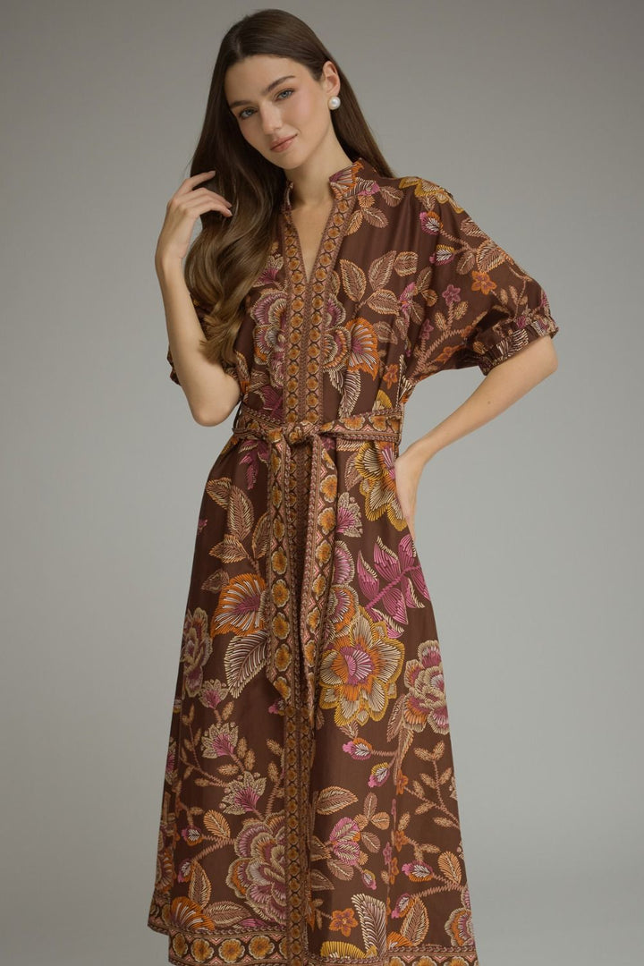 A woman modeling a brown batik maxi dress with a floral pattern in pink, orange, and yellow, featuring a V-neckline, short puff sleeves, and a matching waist tie, set against a plain gray studio background.