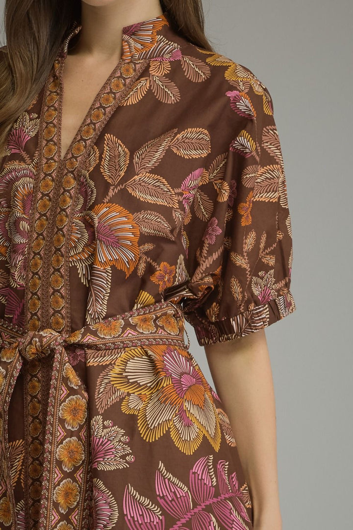A close-up of a model wearing a brown wrap-style dress with a vibrant floral and leaf pattern in orange, pink, and beige, featuring puff sleeves and a matching waist tie.