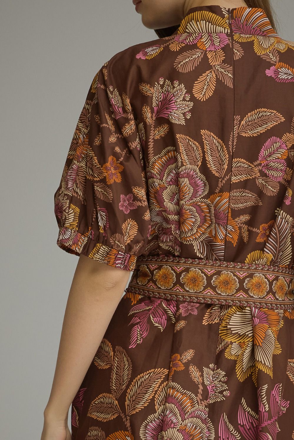A close-up of a model wearing a brown dress with a detailed floral and leaf pattern in pink, orange, and beige, featuring puff sleeves and a matching patterned waistband.